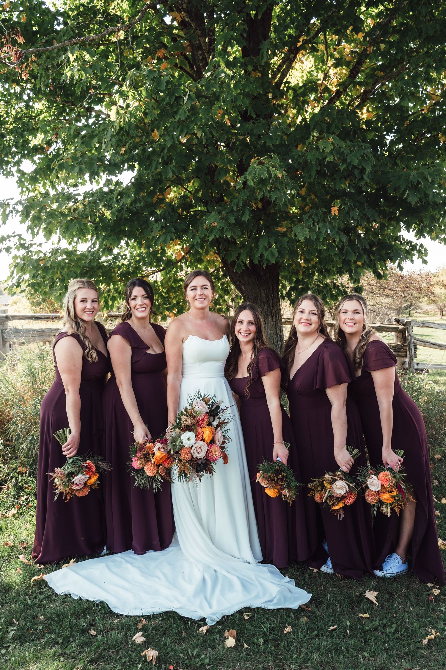 A bridal party poses under a large tree with bridesmaids in burgundy dresses holding autumn bouquets.