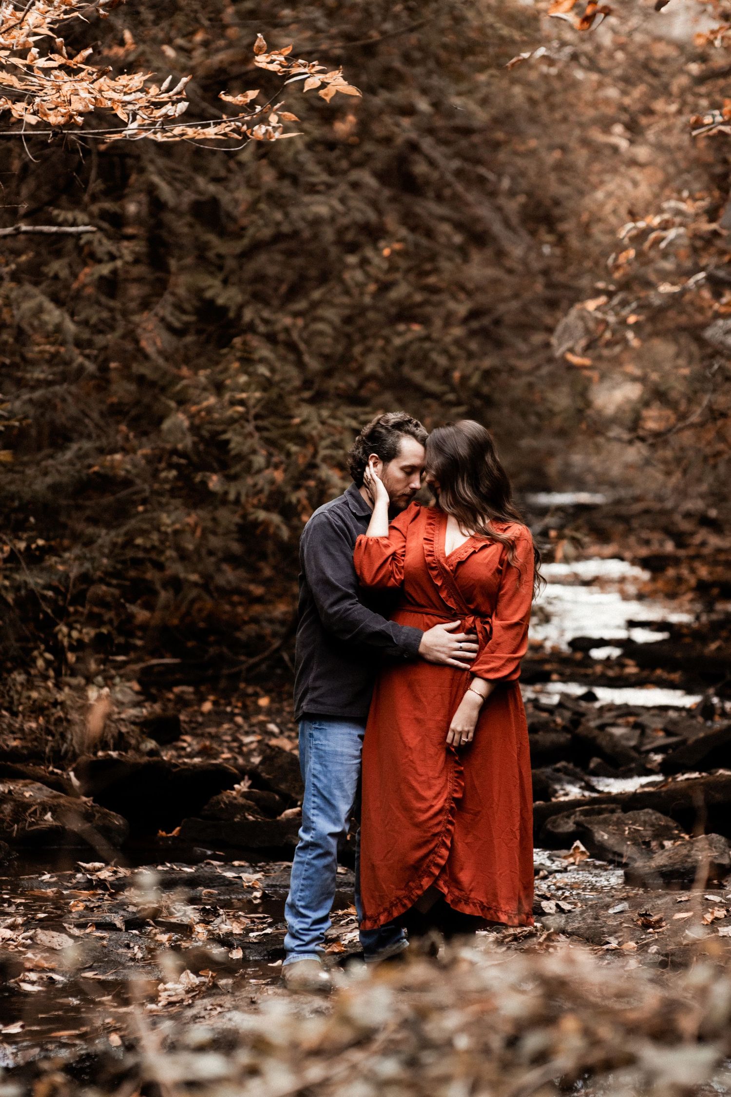 Couple embracing on autumn forest floor with rust dress contrasting against fallen leaves.
