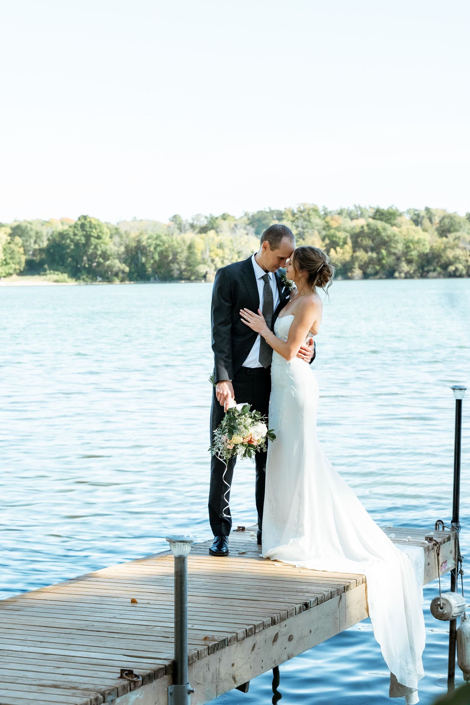 Wedding couple sharing intimate moment on wooden pier with lake and trees in background.