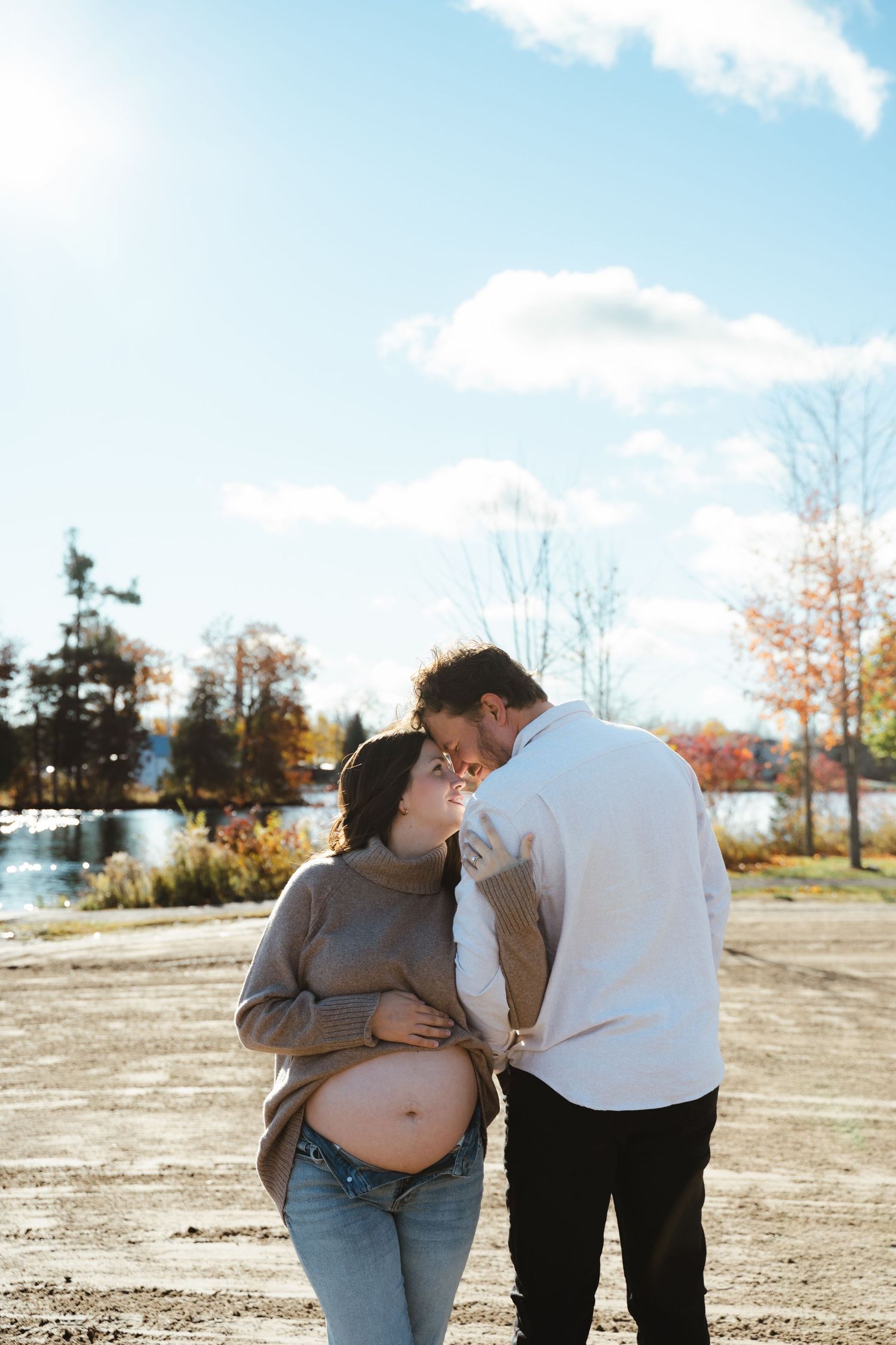 A tender maternity moment outdoors on a sunny autumn day with a scenic lake and trees in the background.
