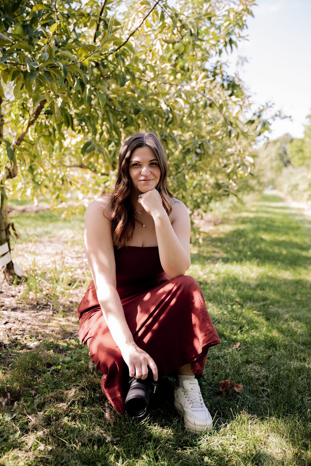 A photographer in burgundy dress poses casually in a sunlit orchard setting.