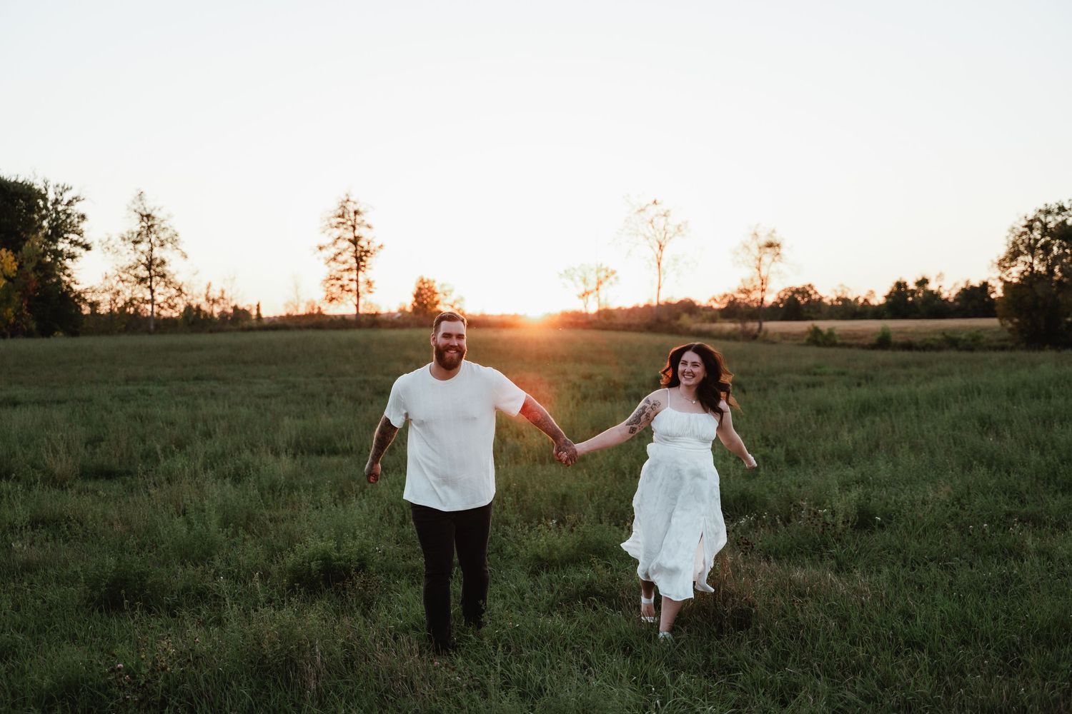 Couple walks hand in hand through grassy field during golden hour photoshoot.