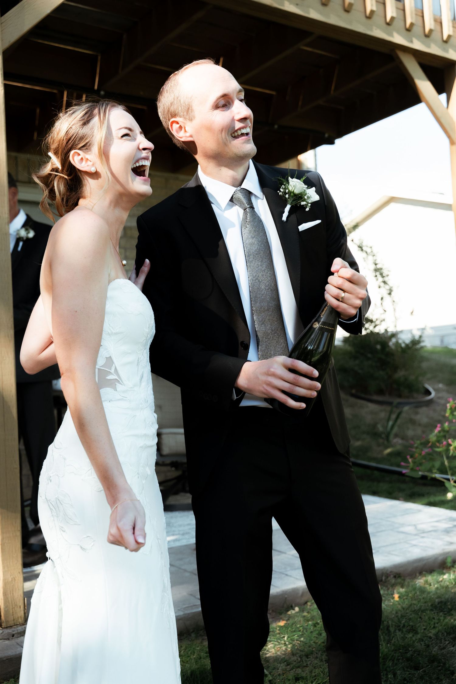 Wedding party laughing together while holding drinks under a wooden pergola.