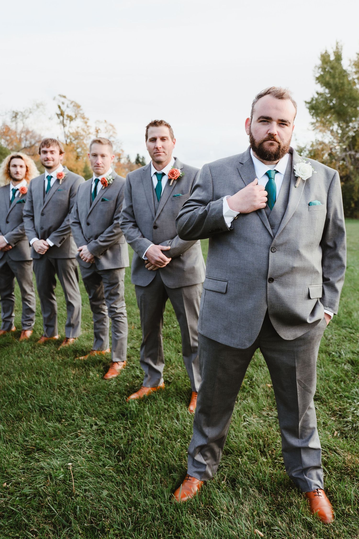 Groomsmen in matching gray suits and teal ties pose on grassy hillside wearing brown leather shoes at Peterborough Curling club
