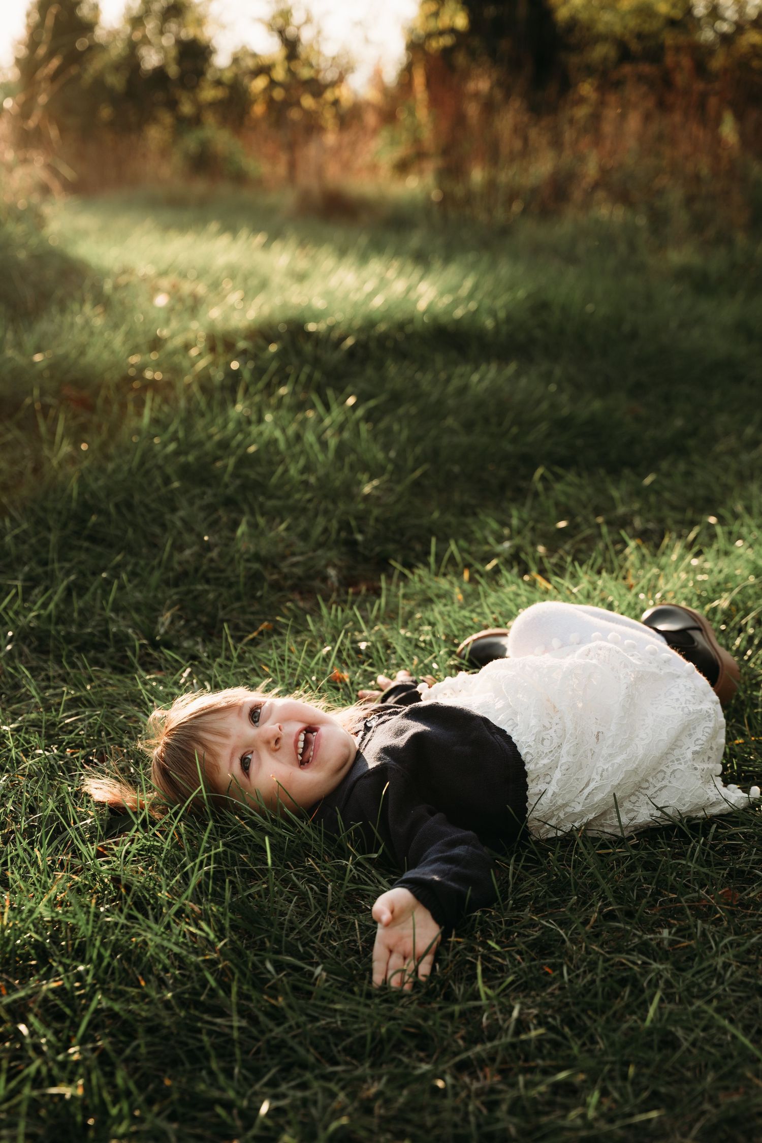 A child in white and black clothing lies laughing in tall grass during sunset.