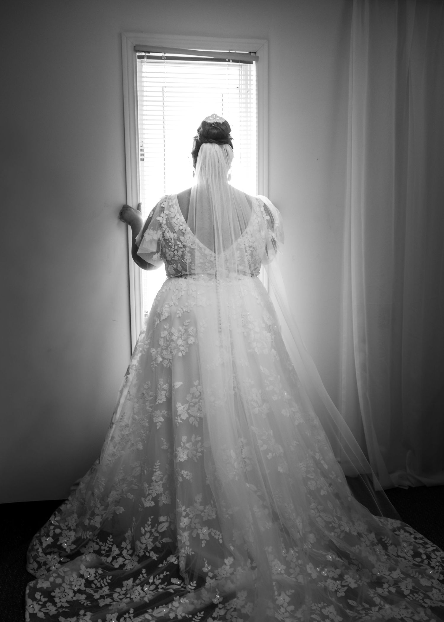 Elegant white lace wedding dress hanging by window with dramatic lighting and flowing train.