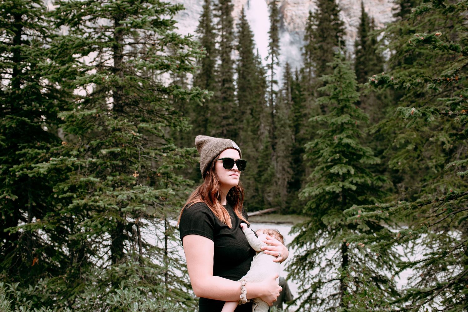 Person in black shirt and beanie standing among tall evergreen trees in forest setting.