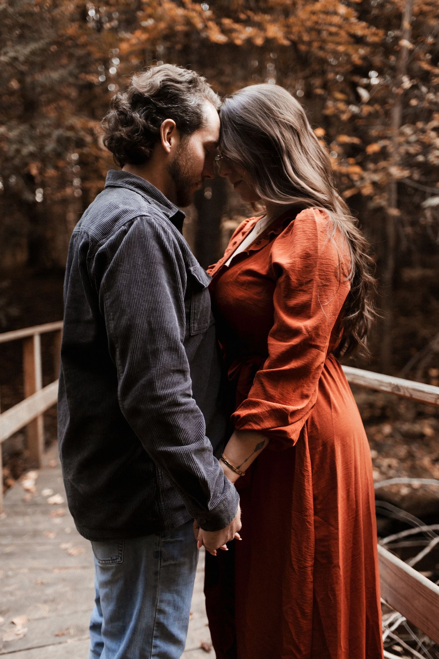 Couple in autumn setting with person wearing flowing rust-colored dress on wooden walkway.