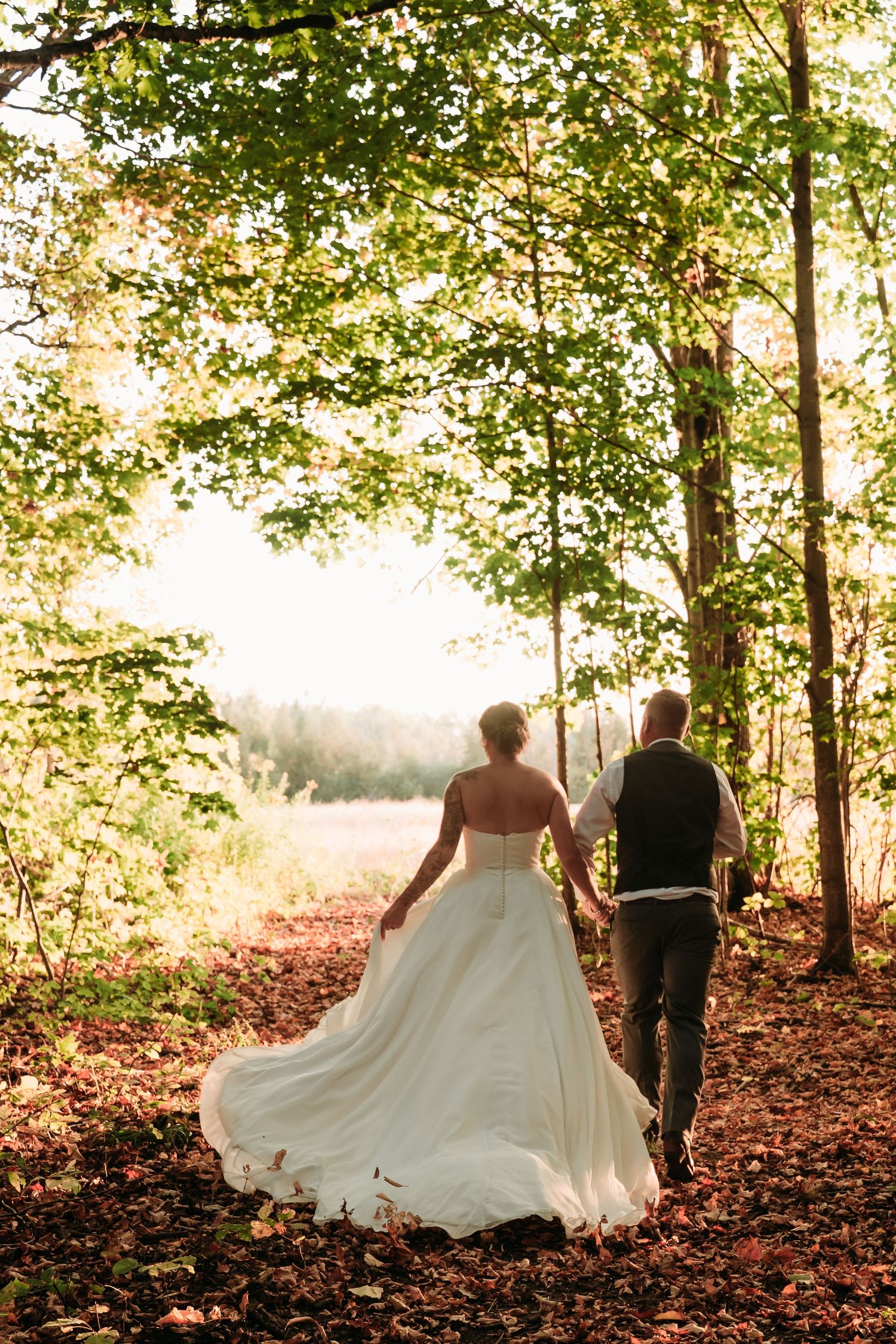 Newlyweds walk hand in hand through a sunlit forest path with flowing white wedding dress.