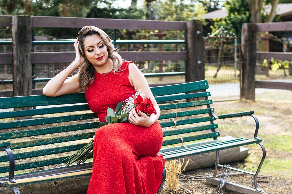 woman in red dress holding red bouquet on a green bench, staring down at bouquet