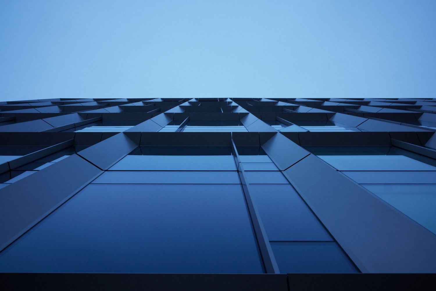 Minimalist architectural facade photographed from below, geometric glass panels rising into a pale blue sky.
