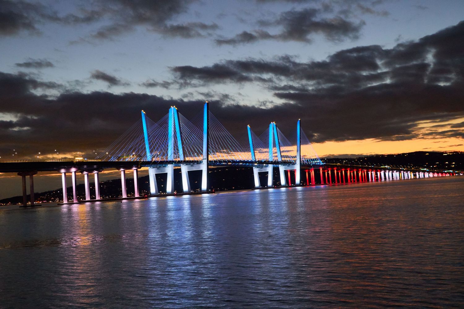 Illuminated cable-stayed bridge spanning the Hudson River at dusk, blue and red lights reflecting across calm water under a dramatic sky.
