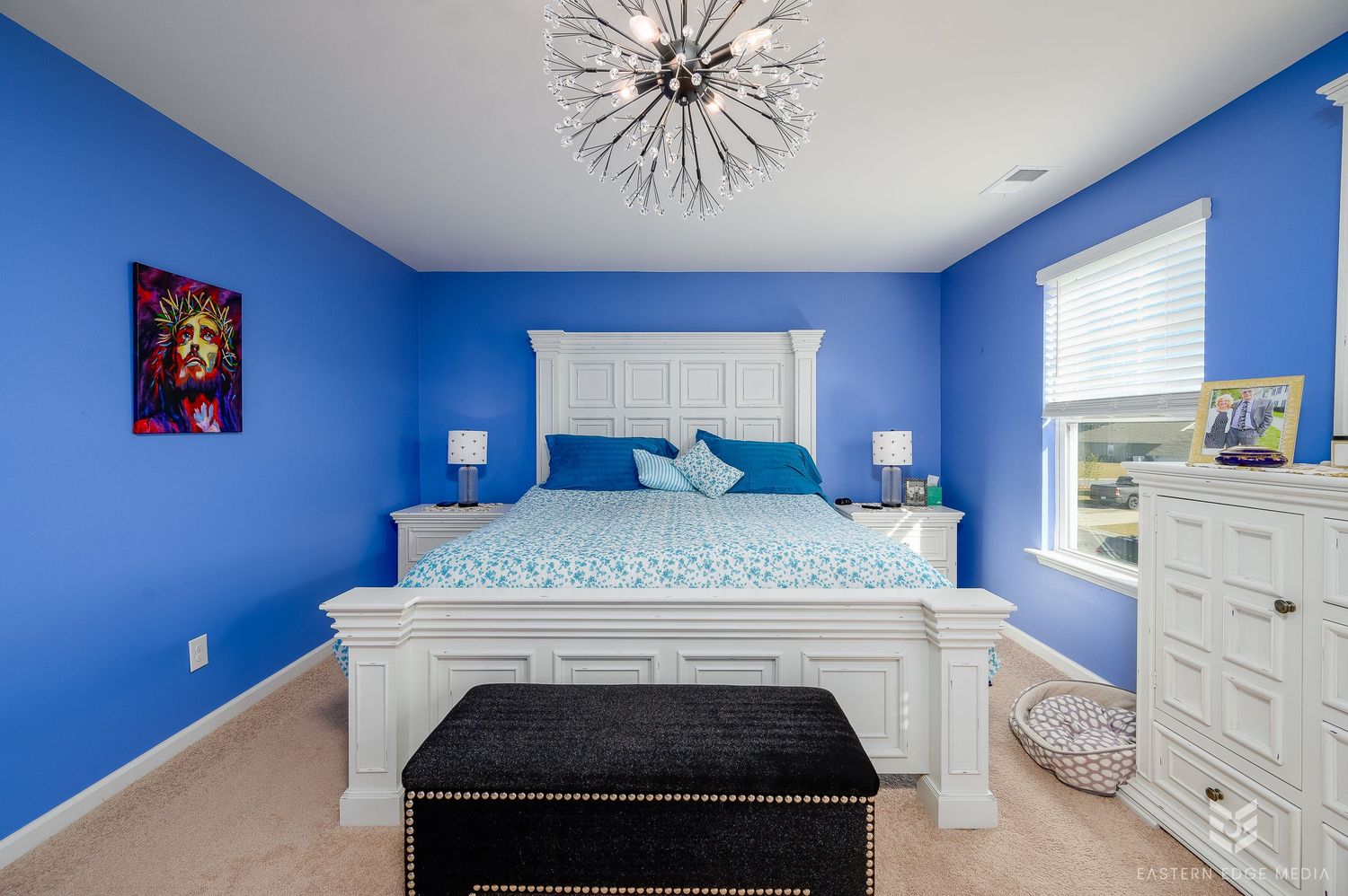 Blue bedroom with white king bed, chandelier, bench, and dresser near window.