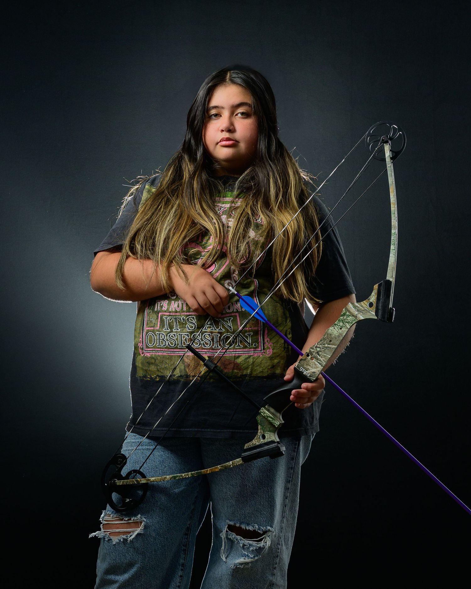 Teen girl poses seriously with compound bow in ripped jeans against dark background.