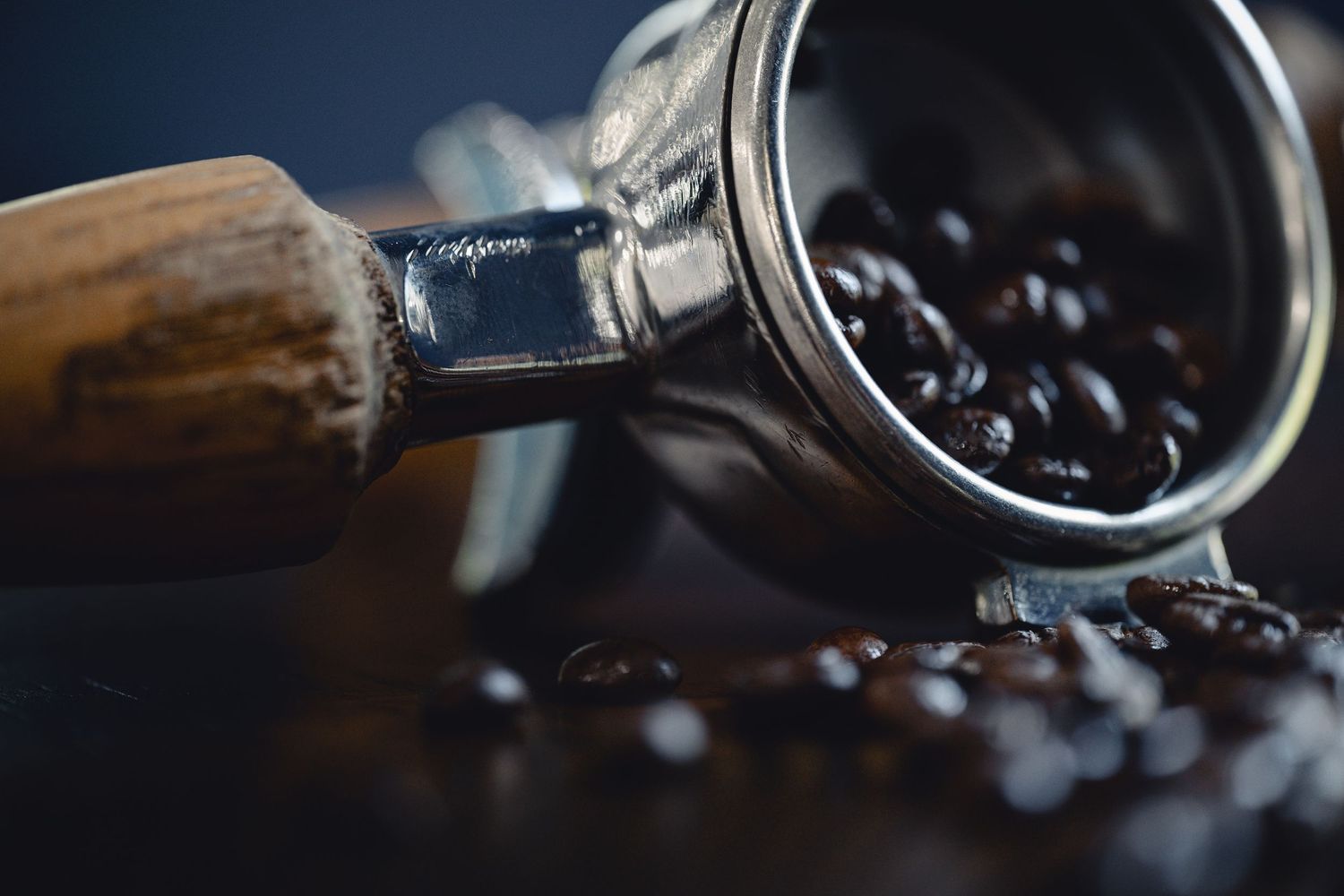 Close-up of wooden-handled espresso portafilter with dark roasted coffee beans on table.