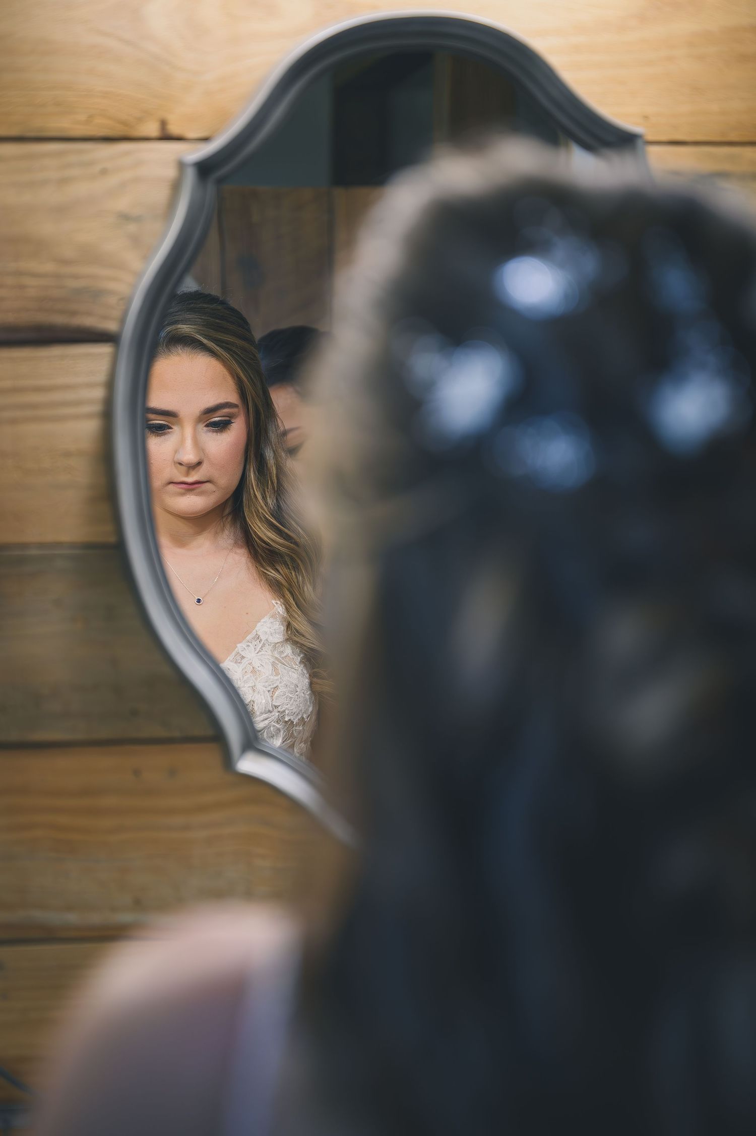 Bride gazing at her reflection in an ornate mirror against a rustic wood wall on her wedding day.