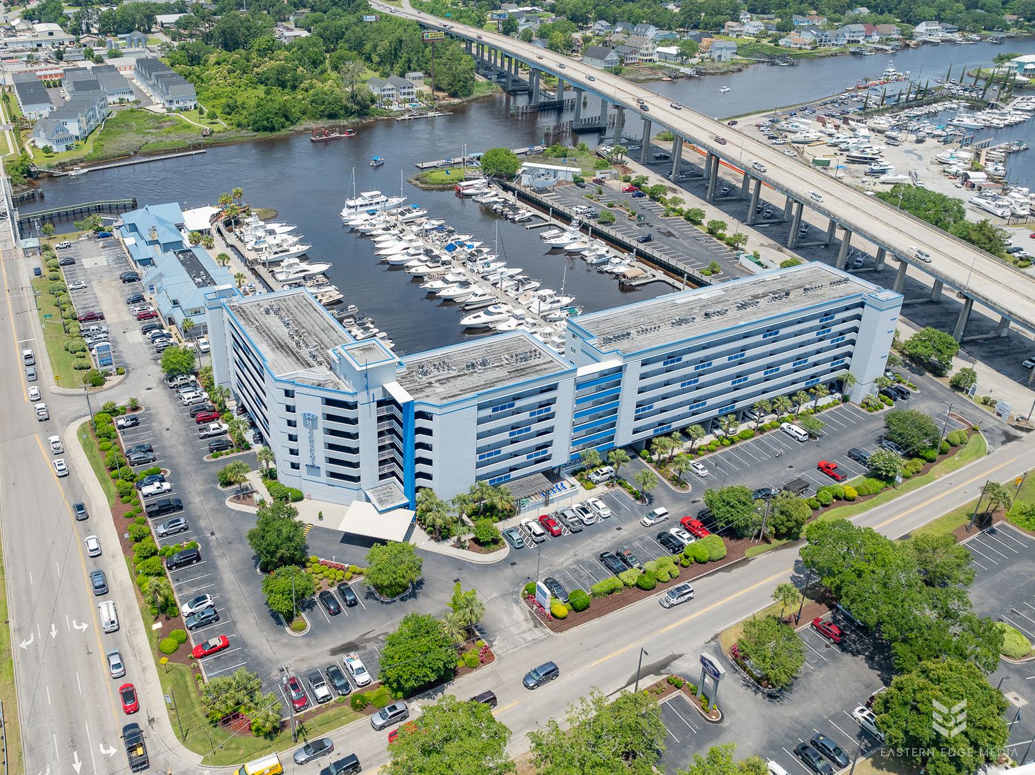 Aerial view of waterfront condominium complex with marina, boats, and bridge nearby.