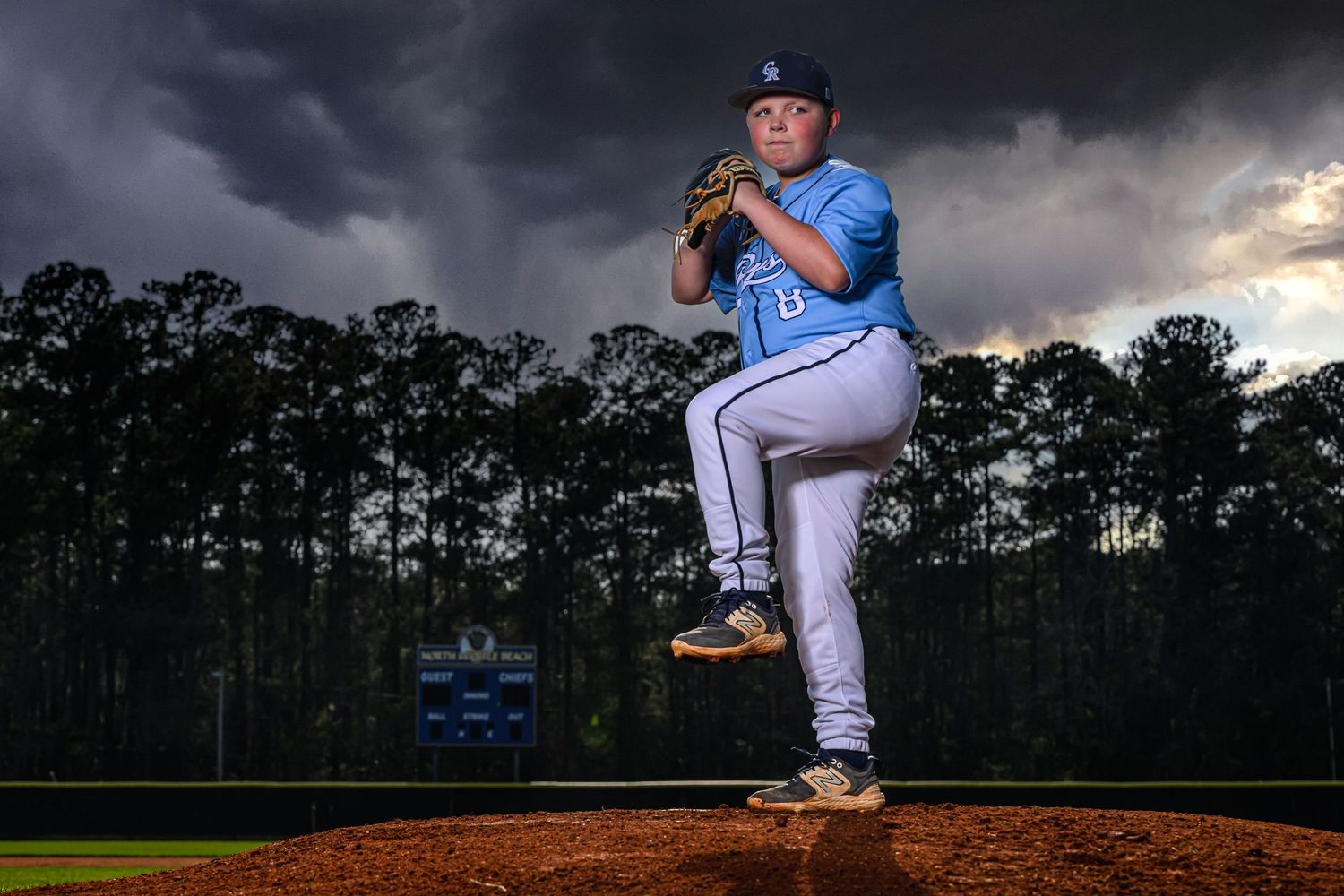 Youth baseball pitcher in a light blue uniform winding up for a pitch on the mound under dramatic storm clouds.