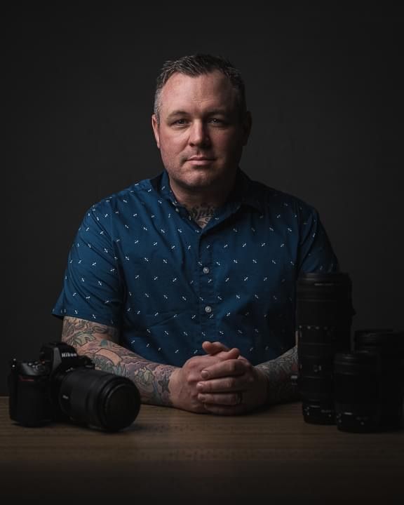 Professional photographer with tattooed arms seated at table with multiple Nikon cameras on dark background.