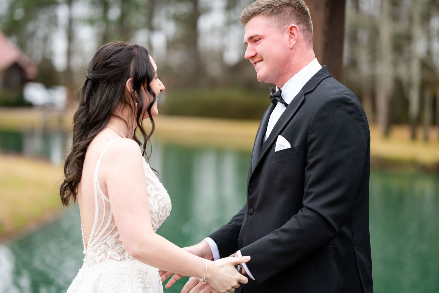 A bride and groom holding hands and smiling at each other during an outdoor lakeside wedding ceremony.