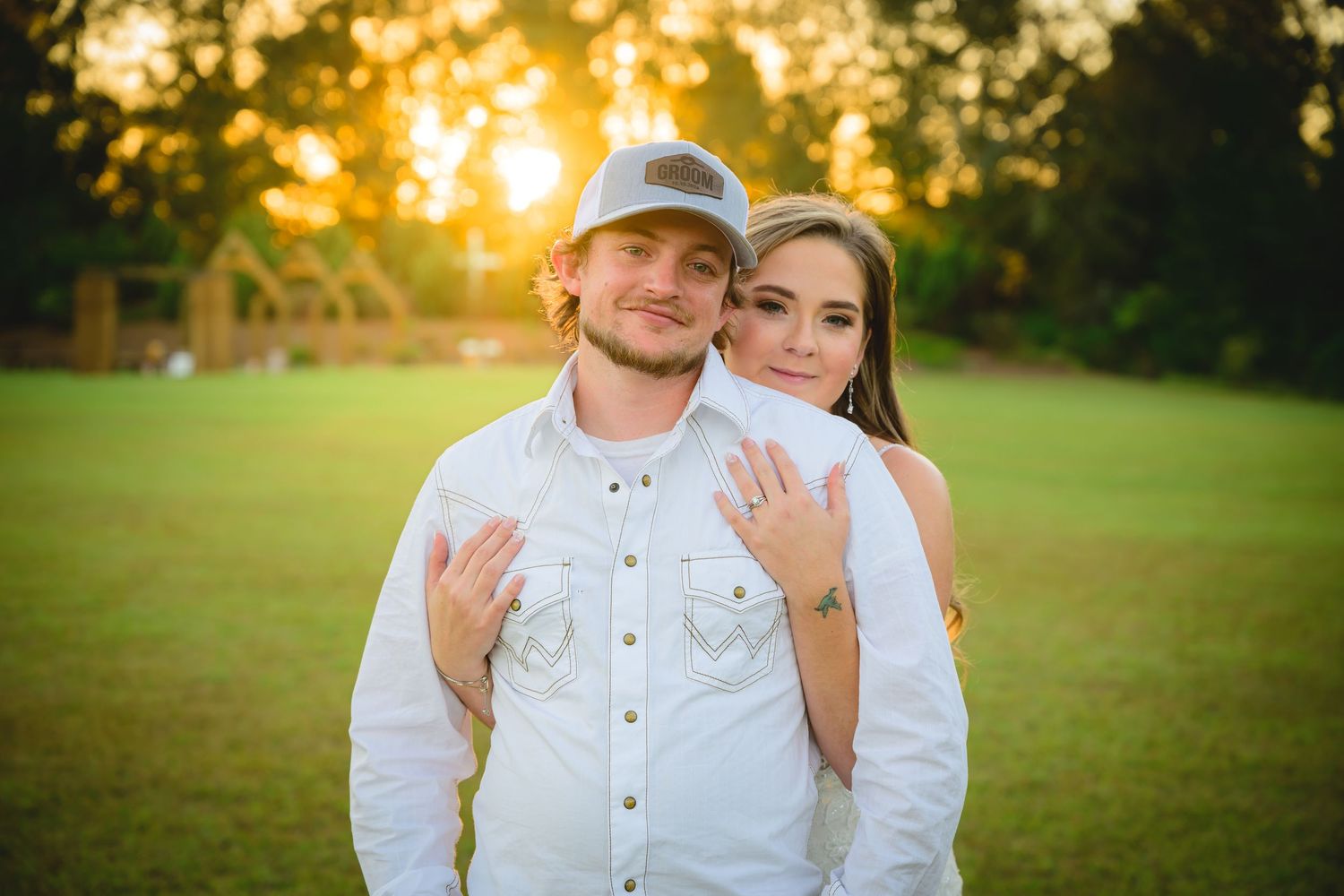 Smiling bride and groom posing together at golden hour sunset in an open green field.