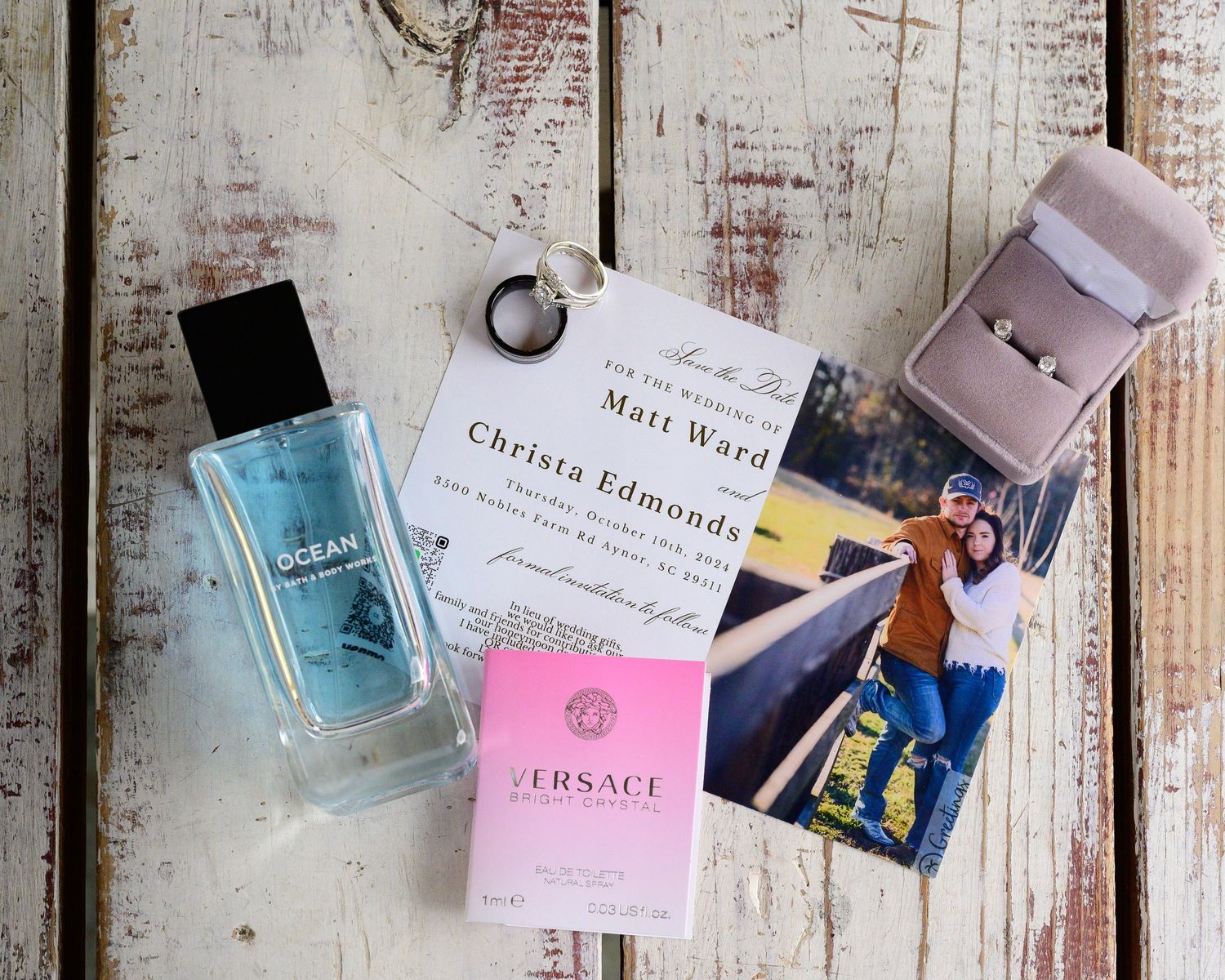 Flat lay of wedding invitation, Versace perfume, ring box, and couple photo on rustic wood.