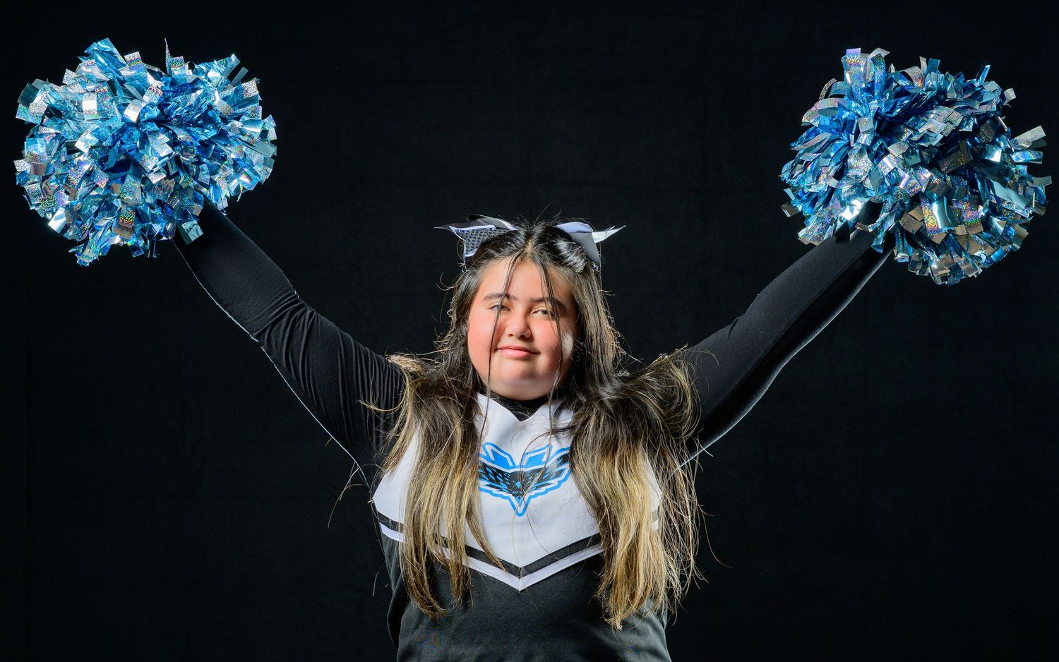 Young cheerleader in uniform holds blue pom-poms overhead against dark background.