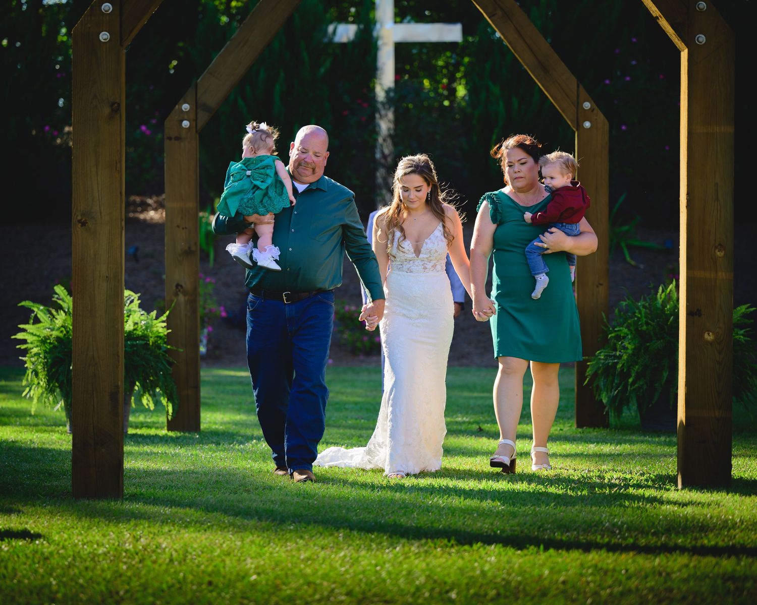 Wedding party walking through a wooden arch on green lawn with family carrying young children.