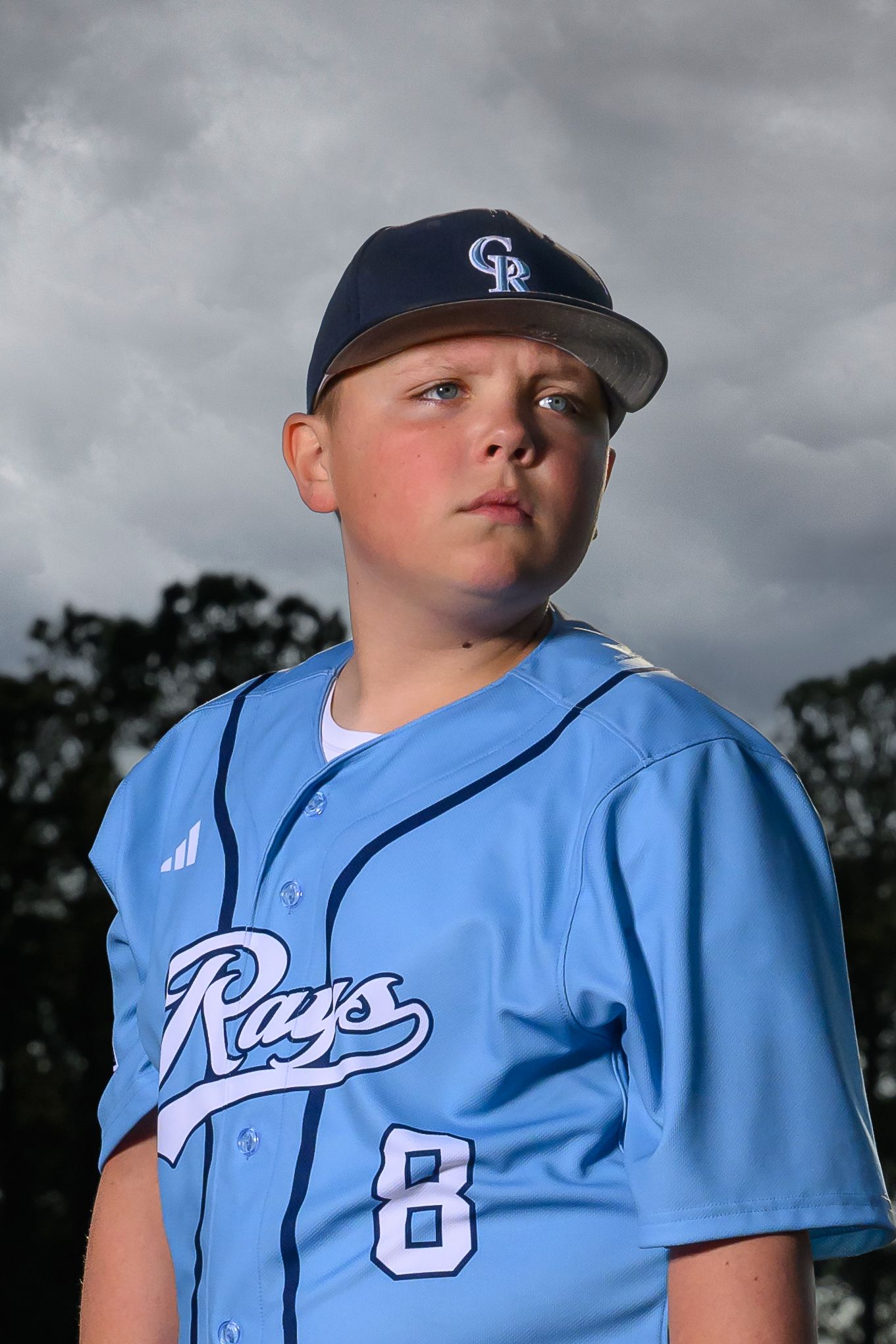 Young baseball player in a light blue Rays jersey number 8 posing for a portrait under cloudy skies.