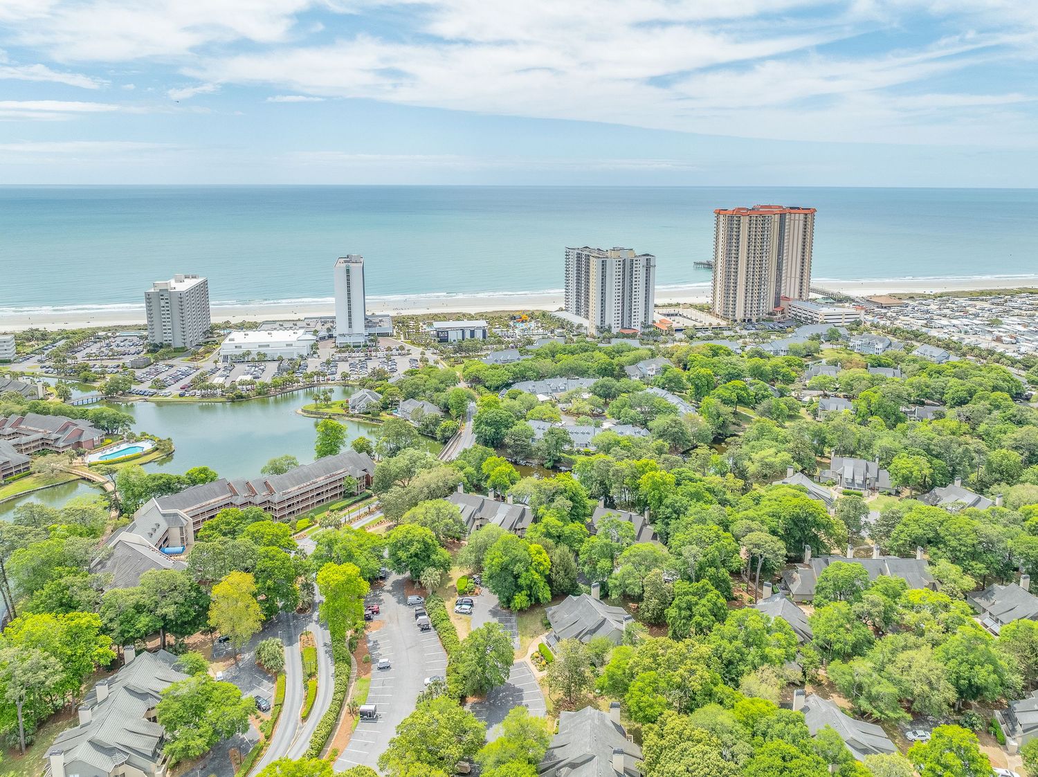 Aerial view of a coastal town with high-rise buildings, lush greenery, a lake, and the ocean in the background.