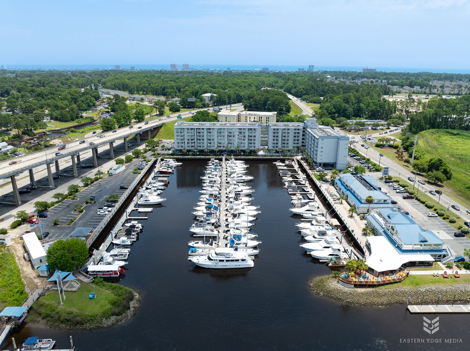 Aerial view of marina with boats, waterfront hotel, and lush surrounding landscape.