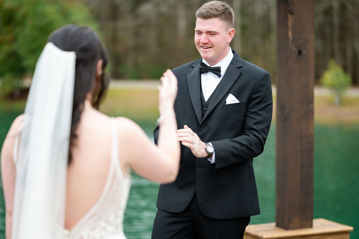 A groom in a black tuxedo smiling while holding his bride's hands during an outdoor wedding ceremony.