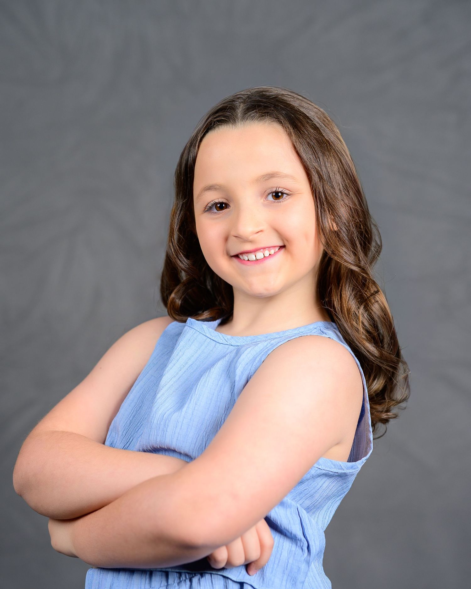 Smiling young girl in blue dress poses with arms crossed in studio portrait.
