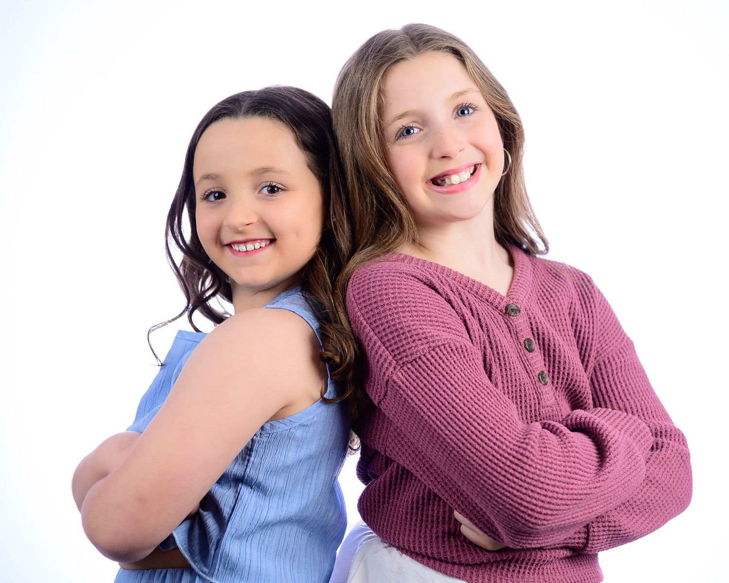 Two young girls smile back-to-back in coordinated studio portrait on white background.