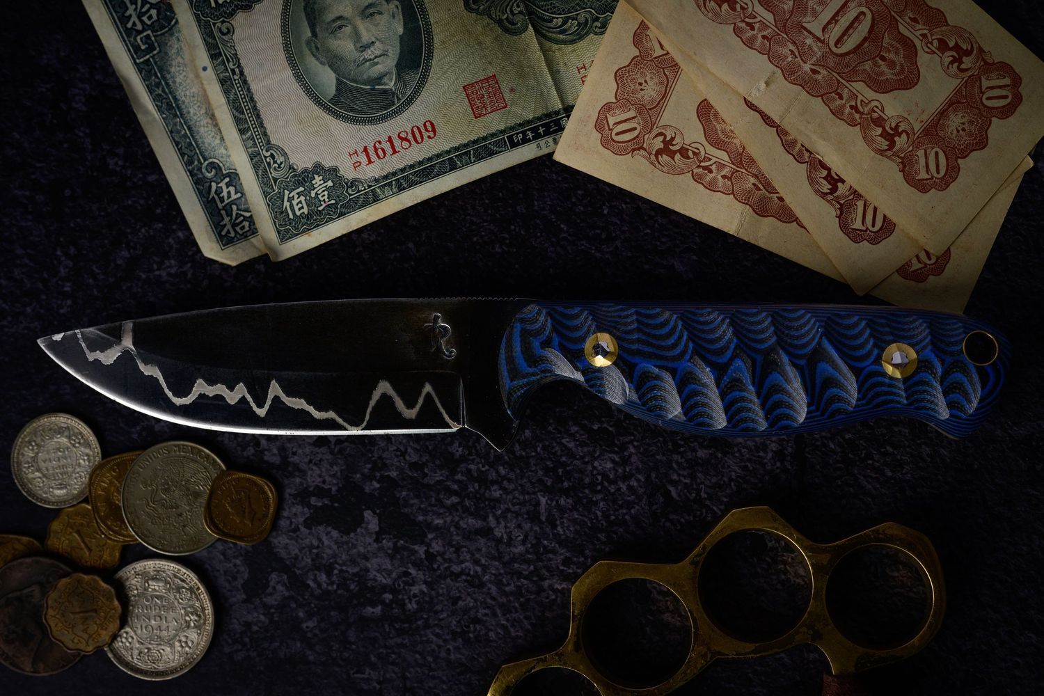 A tactical folding knife with blue handle displayed alongside foreign currency, coins, and brass knuckles.