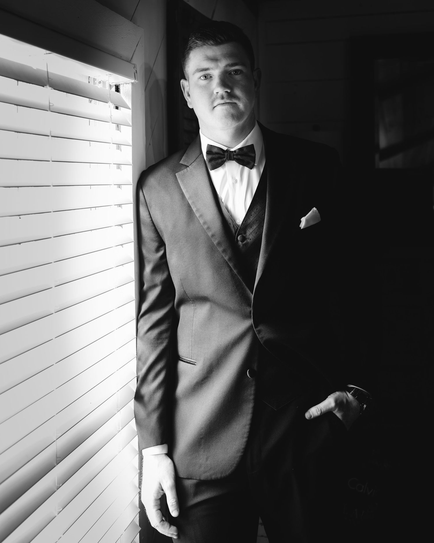 A black and white portrait of a groom in a tuxedo standing by a window with venetian blinds.