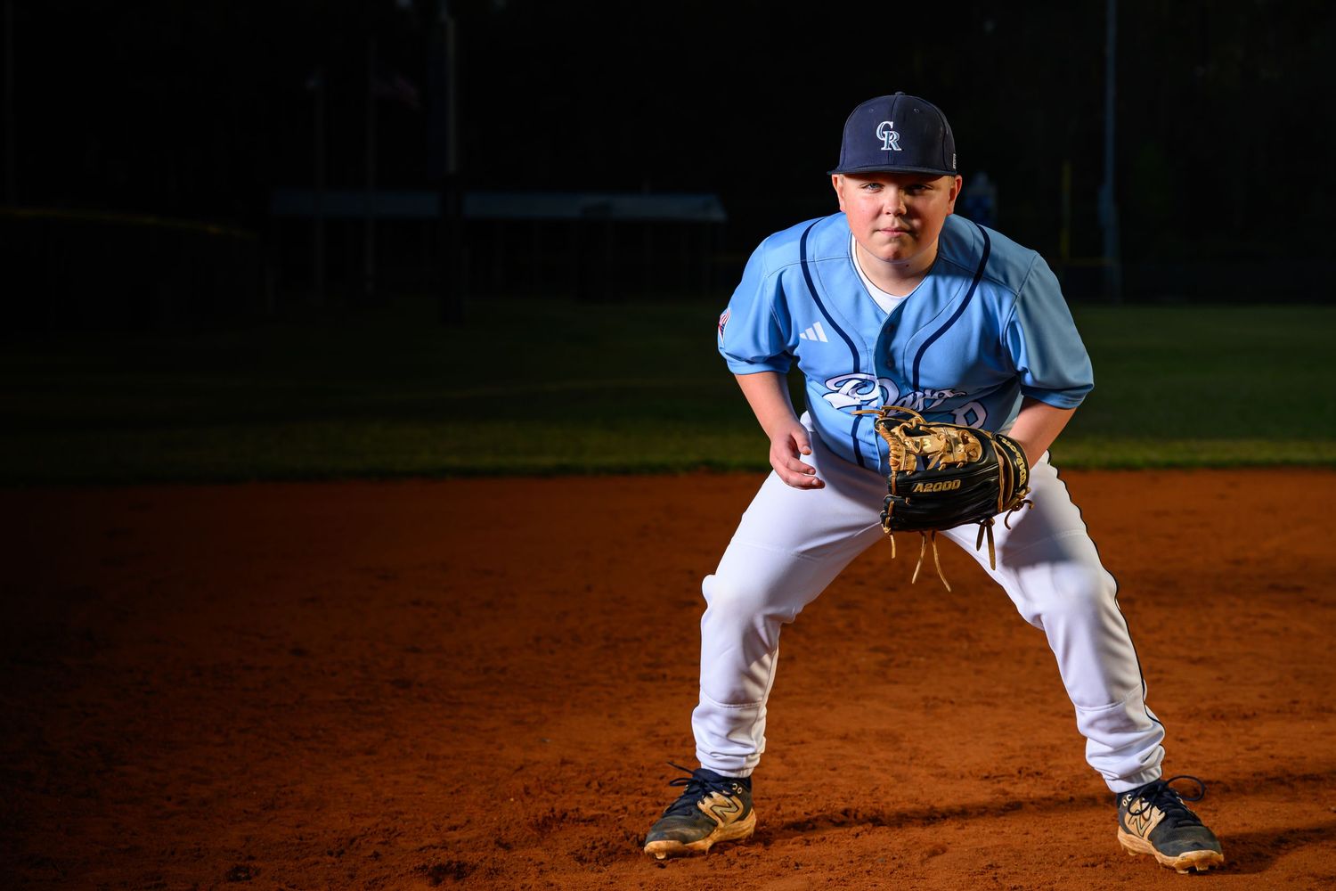 Young baseball player in a light blue uniform in a ready fielding stance on a dirt infield at night.