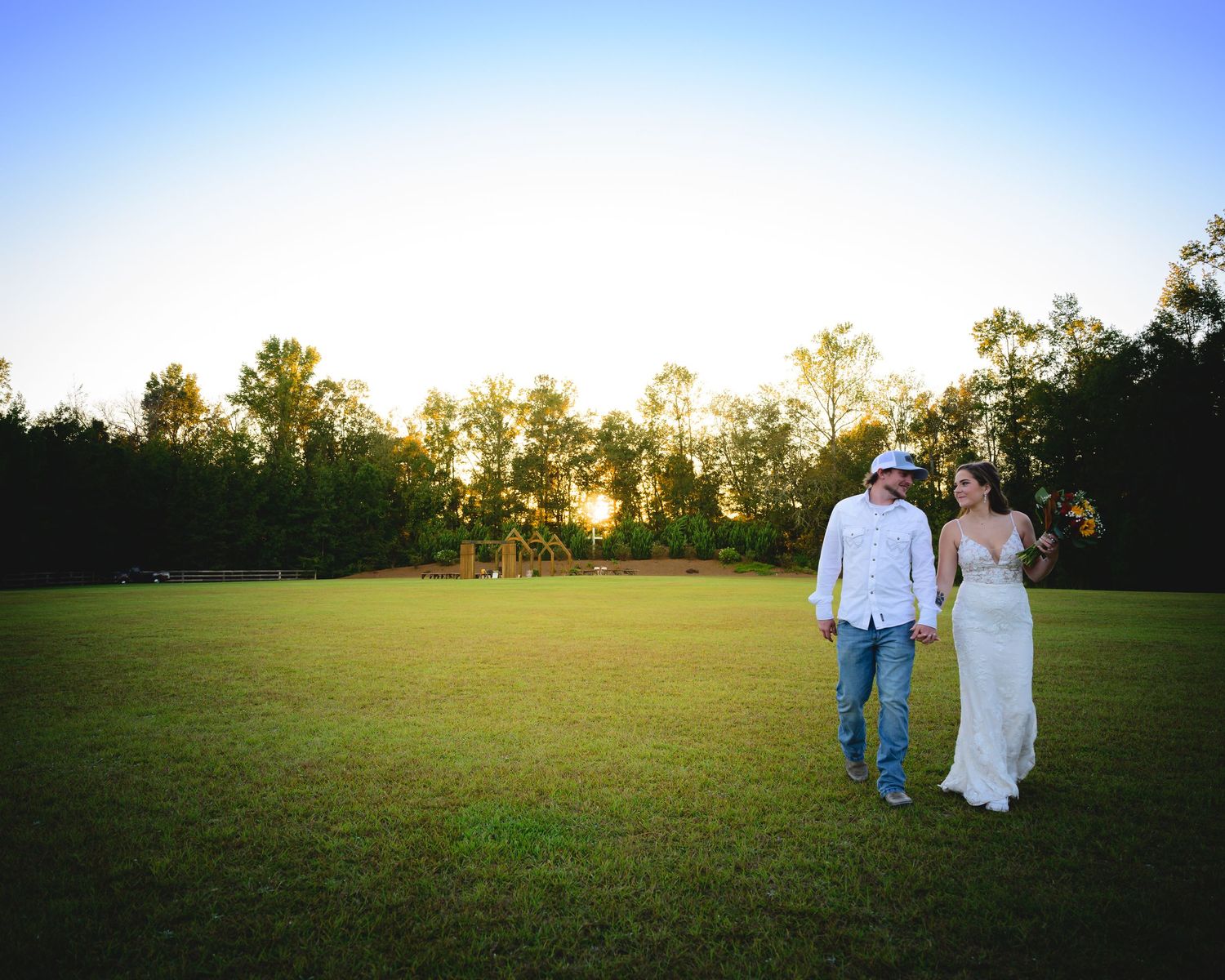 Bride and groom walking hand in hand across a lush green field at a beautiful sunset.