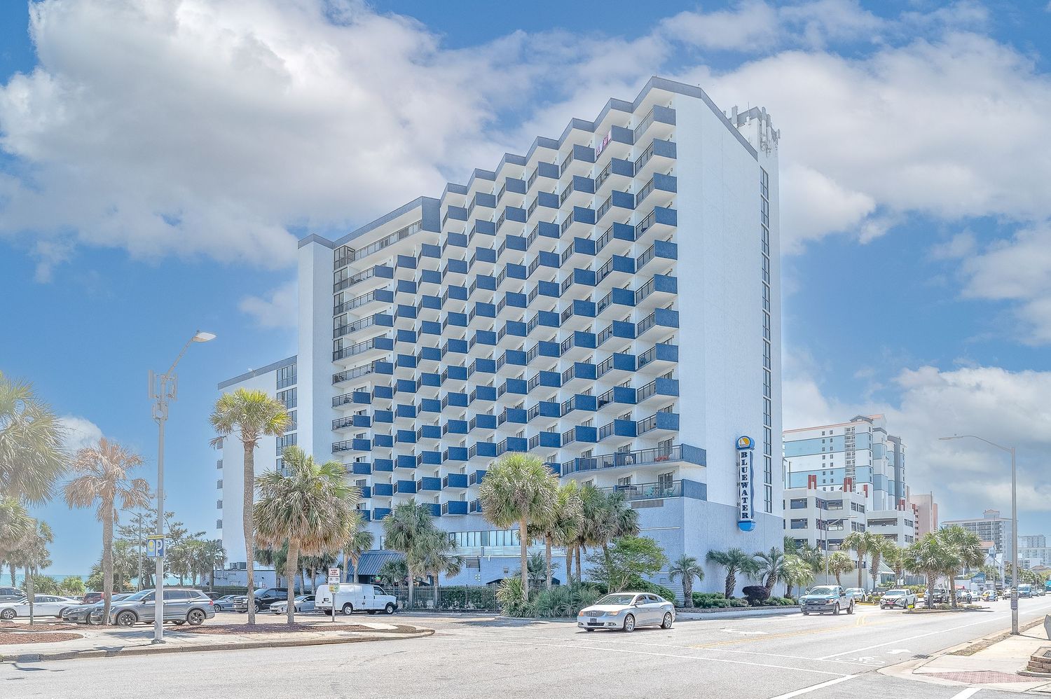 Large white oceanfront hotel with multiple balconies and palm trees under a bright blue sky.