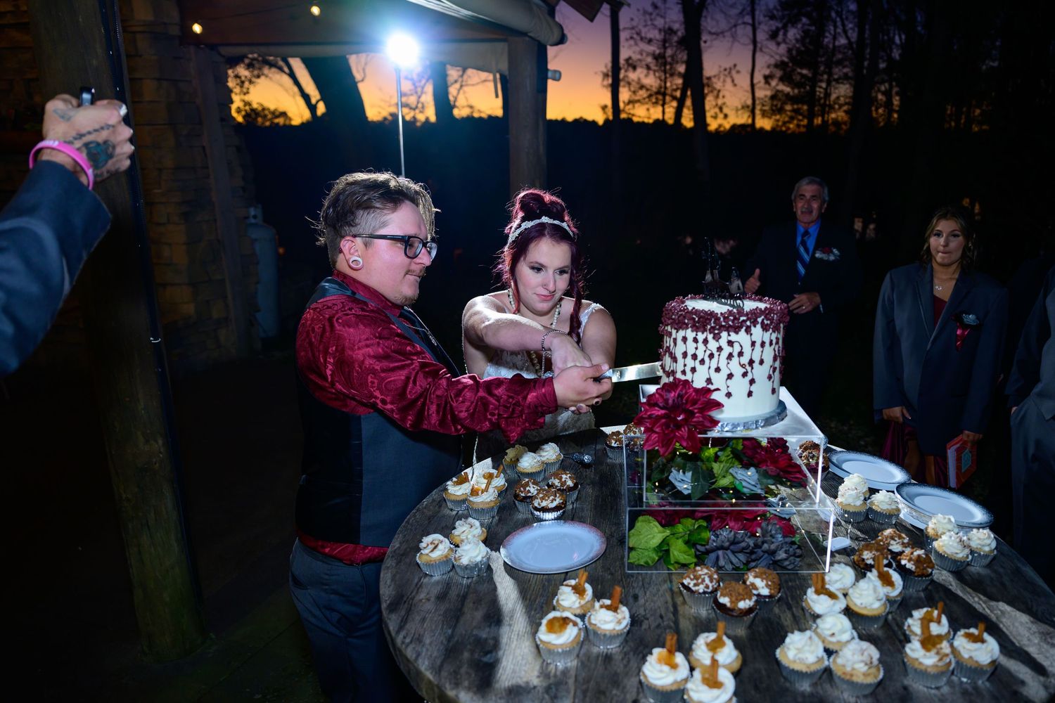 Bride and groom cut wedding cake at outdoor evening reception with dessert table at sunset.