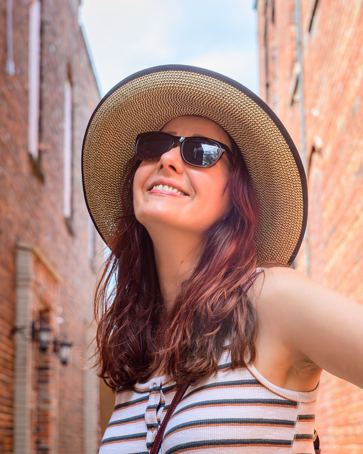 Smiling young woman wearing a straw sun hat and sunglasses taking a selfie in a brick alleyway.