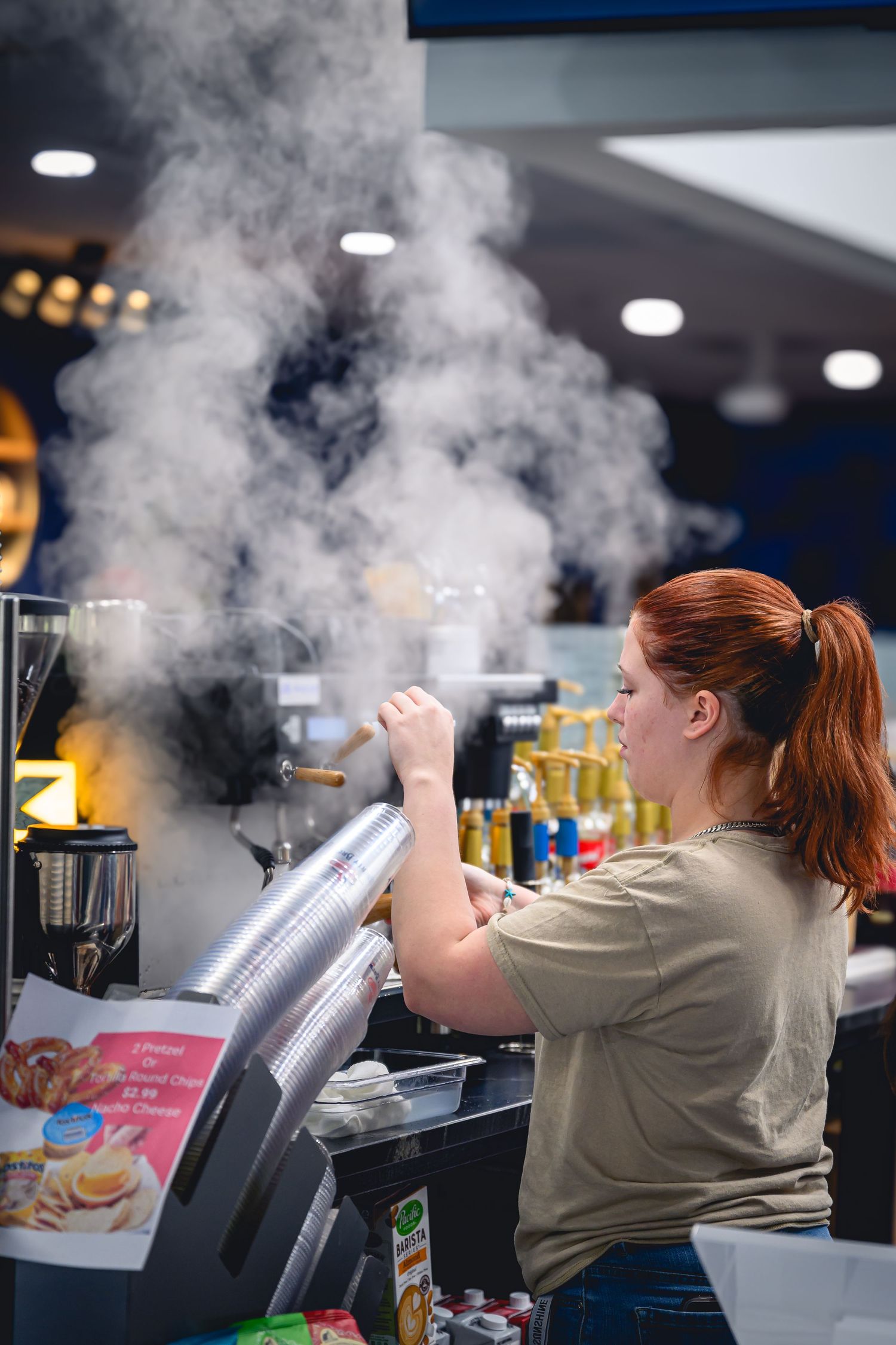 Red-haired female barista operating steam wand on espresso machine in busy coffee shop.