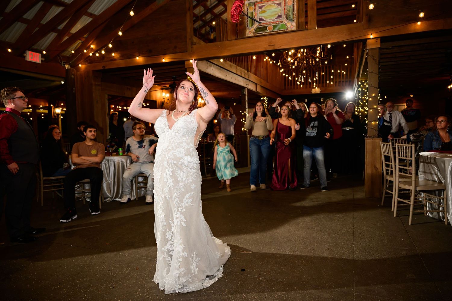 Bride in white lace gown tossing bouquet at wedding reception in rustic barn venue with string lights.