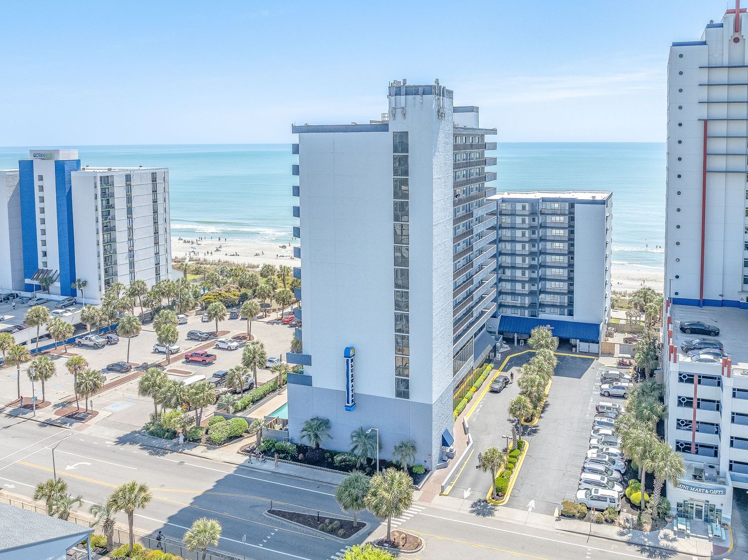 Aerial view of a beachfront hotel tower near the ocean surrounded by coastal buildings and parking lots.