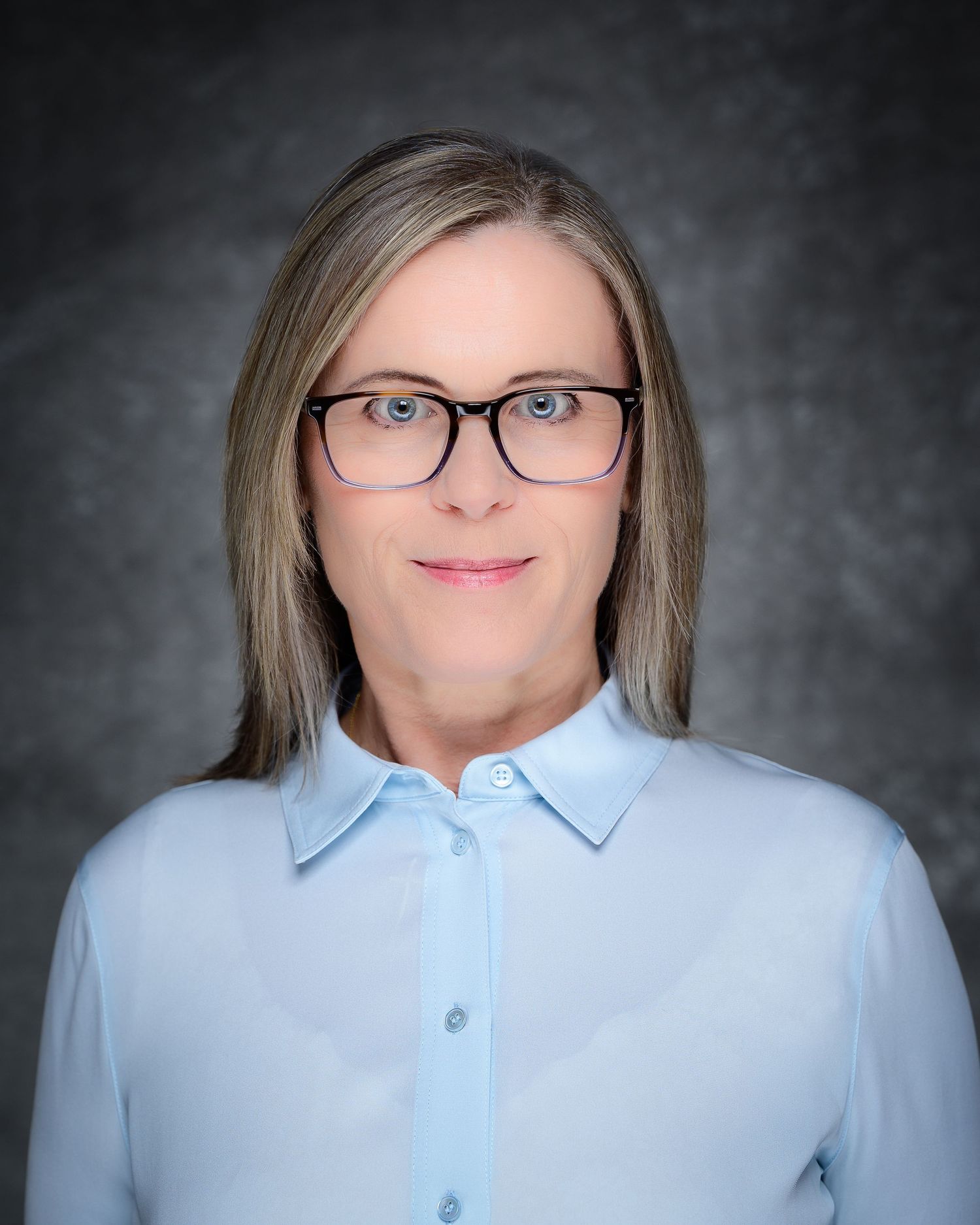 Professional headshot of a woman with straight blonde hair and dark-rimmed glasses in a light blue blouse.
