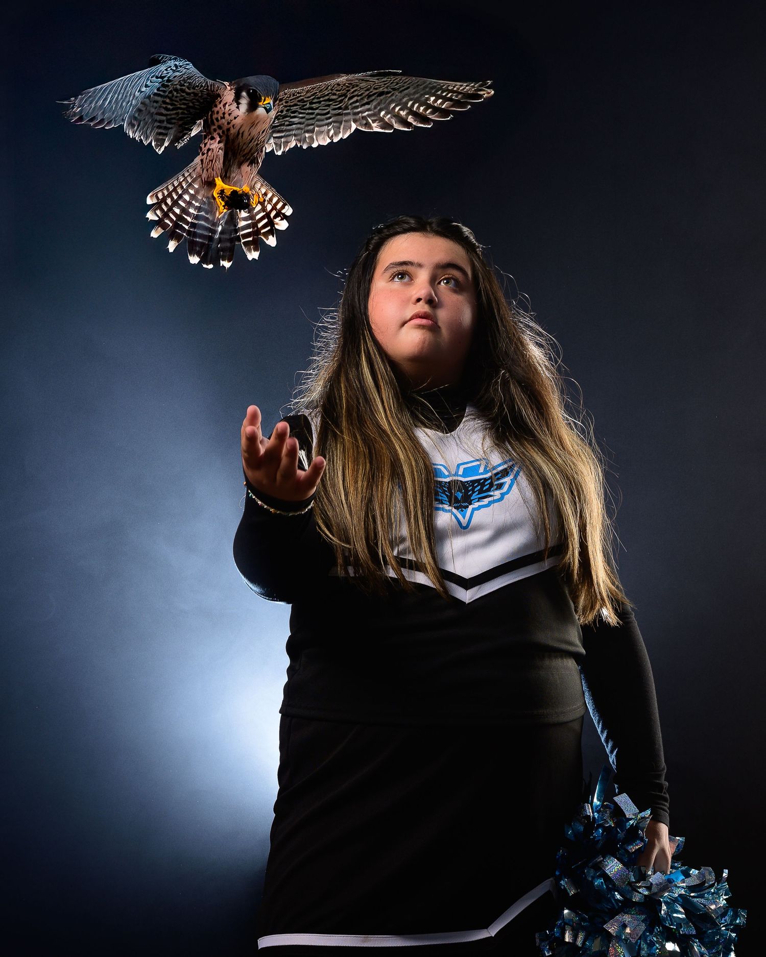 Cheerleader in black uniform reaches up toward a hawk in flight against a dramatic dark blue background.