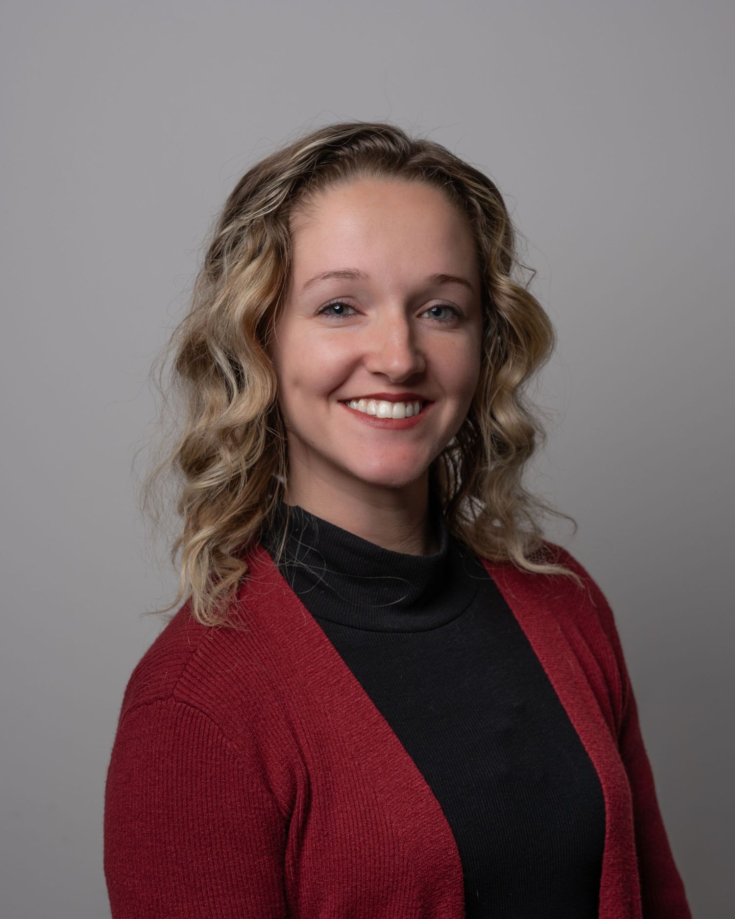 Smiling young woman with curly blonde hair in red cardigan poses for headshot.