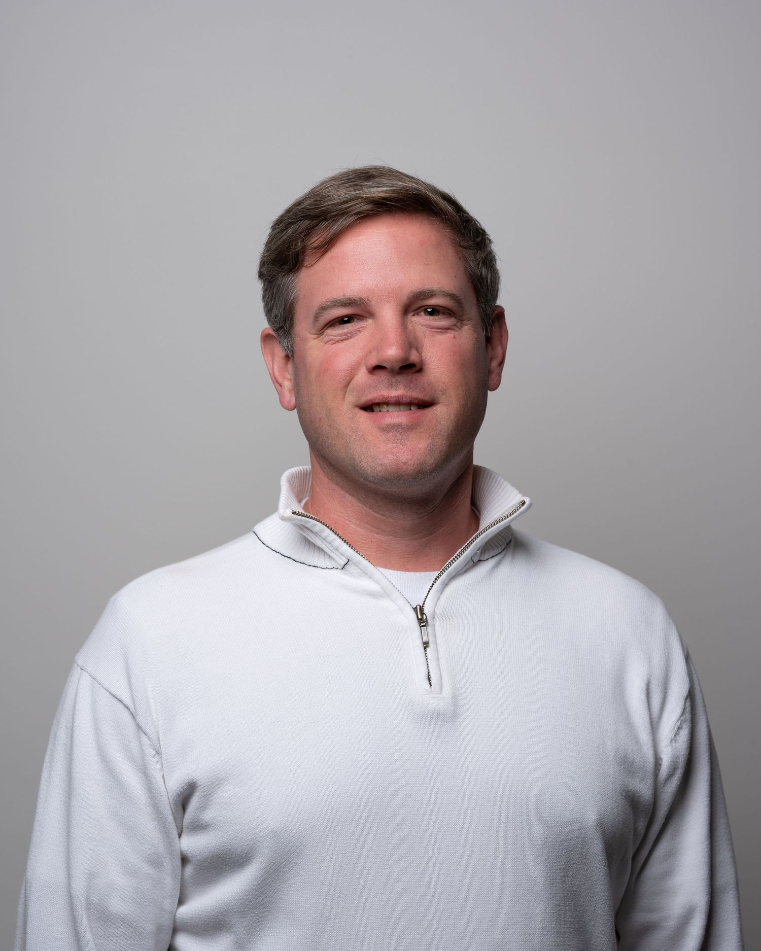 Smiling man in white quarter-zip pullover poses for professional headshot on gray background.