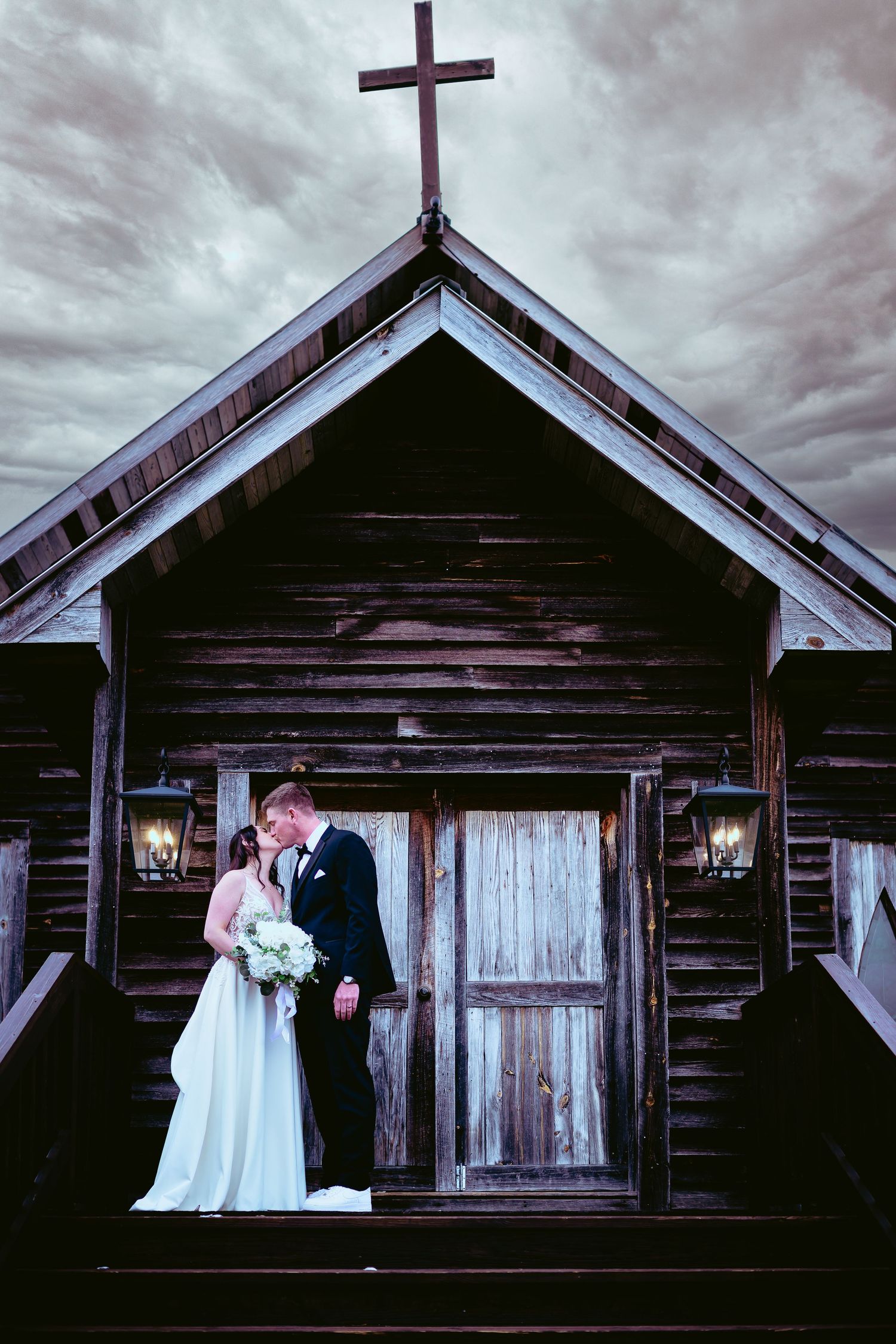 Bride and groom pose romantically outside rustic wooden chapel with cross under dramatic sky.