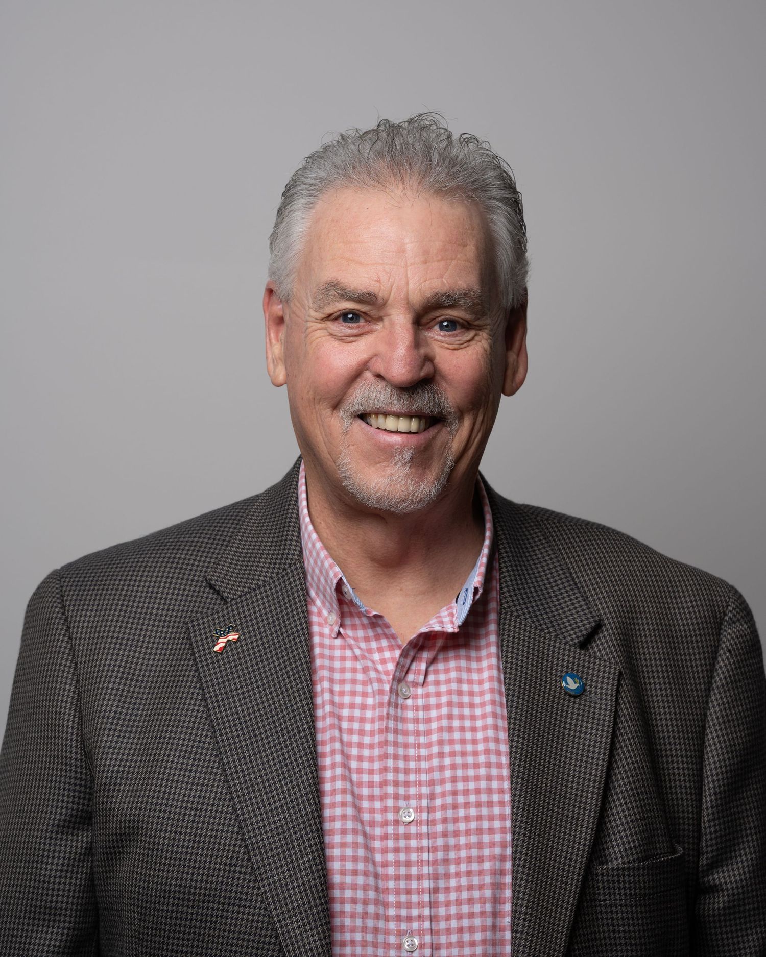 Smiling senior man with gray hair and beard wearing a pink checkered shirt and gray blazer against a gray background.
